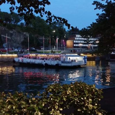 Fullt med människor ombord på restaurangfärjan Jakke jokilautta. Färjan lyser i rött och vitt i ån då han tar i land vid Teaterbron i Åbo en sommarnatt.