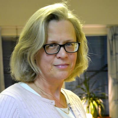 en kvinna i ljust hår och ljusa kläder och mörka glasögon står mitt i ett kontorsrum