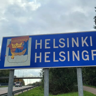 En vägskylt där det står Helsinki Helsingfors och där stadens vapen med en båt och en krona syns, men det är delvis täckt med rödgul tejp.