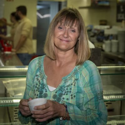 En medelålders kvinna med blont går och blå tröja står i en butik. I händerna håller hon en kaffekopp.