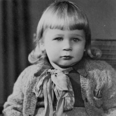Vanhassa valokuvassa pikkulapsi rusetti kaulassaan ja neuletakissaan katsoo totisena kameraan. Lapsella on otsatukka ja olkapäille kihartuvat hiukset.