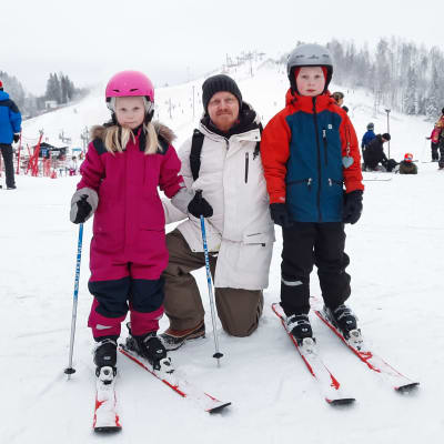 En man och två barn med slalomskidor poserar för kameran framför en slalombacke.