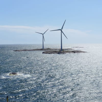 Tre vindkraft stående på klippor ute i ett soldränkt hav.