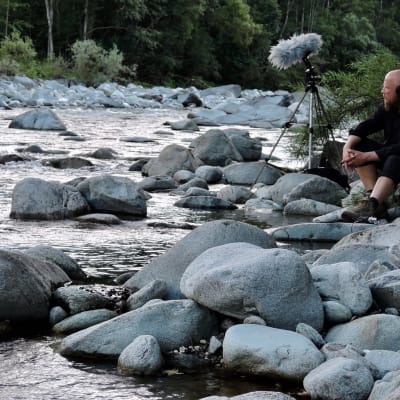 Mies istuu kiven päällä virtaavan veden äärellä. Hänen vierellään näkyy mikrofoni, jolla hän taltioi ympäristön ääniä.