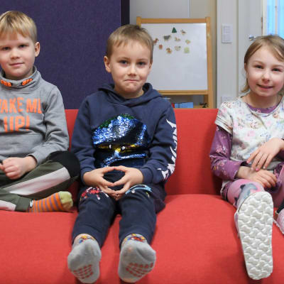 Bilden föreställer tre glada förskolebarn som sitter i en röd soffa.