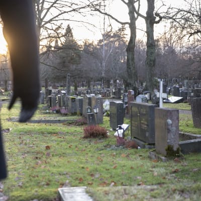Mies on hautausmaalla ja katselee hautakiviä. Hän seisoo kuvan vasemmassa laidassa ja hänestä näkyy vain osa vartaloa ja oikea käsi. Laskeva talvinen aurinko pilkottaa käden kainalosta.