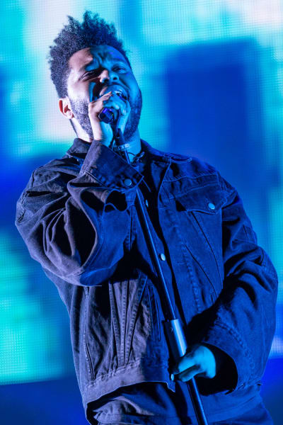 The Weeknd håller i en mikrofon och sjunger på scen. Han är klädd i svarta kläder från topp till tå. I bakgrunden en blå skärm.