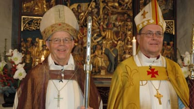 Biskop Gustav Björkstrand och ärkebiskop Jukka Paarma klädda i biskopsskrudar och med mitra och stav sida vid sida framför altaret i Pernå kyrka.