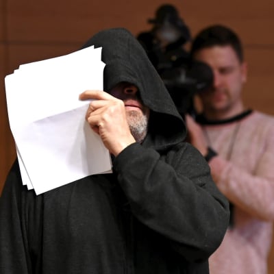 Mika Moring gömmer sitt ansikte med en huva och en pappersbunt på sin rättegång 23.12.2022.