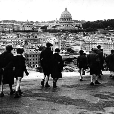 Joukko nuoria poikia kävelee kadulla poispäin kamerasta, taustalla näkyy Rooman kaupunkia ja Pietarinkirkon kupoli. Kuva elokuvasta Rooma - avoin kaupunki.