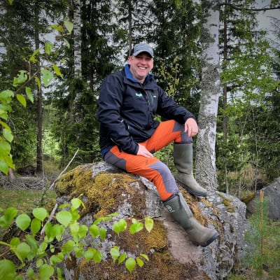 En man sitter på en sten i skogen och ler.