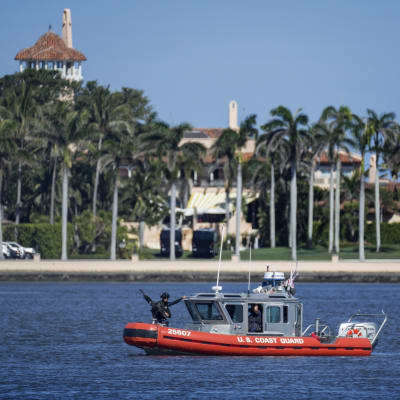 Kustbevakningens lilla röda båt med beväpnade vakter skyddar strandlilnjen till rpesidnetens lyxklubb som skymtar bakom palmerna.