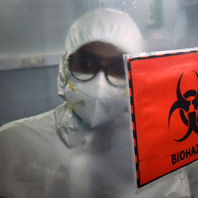 En person i skyddsoverall och munskydd står bakom en varningsskylt där det står "biohazard".