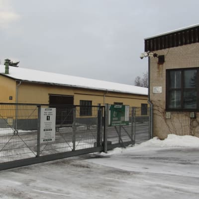 Två fabriksbyggnader och ett staket av metallnät. Stängd port till området. Snö och vinter.