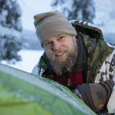 Hymyilevä Mikko Kettunen pystyttää vihreää telttaa lumisessa metsässä.