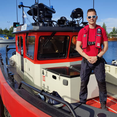 Mies seisoo pelastusveneen kannella kesähelteellä. Taustalla näkyy merta.