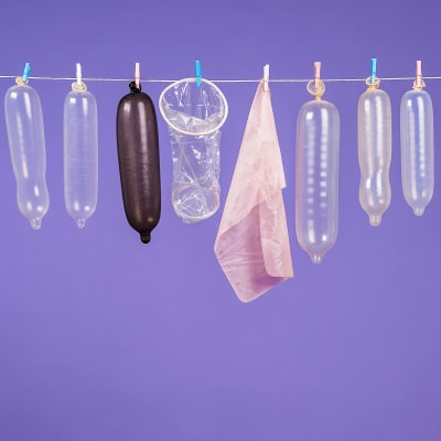 Erilaisia ja eri värisiä kondomeja sekä suuseksisuoja roikkuu pyykkinarulla violettia taustaa vasten.