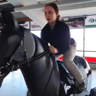 nainen ratsastaa simulaattorilla, valmentaja katsoo vierestä