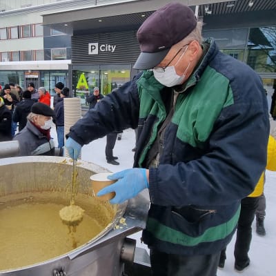 En äldre man skopar ärtsoppa ur stort soppkök. I bakgrunden en liten folkhop, många i reflexvästar.