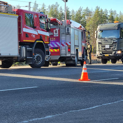Militärfordon och brandbilar vid en olycksplats vid en landsväg. Det fins orangea koner utplacerade på vägbanan för att dirigera om trafiken.