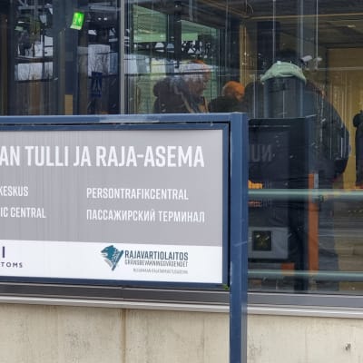 På en skylt utanför en byggnad står det Nuijamaa tull och gränsstation - persontrafikcentral på finska, svenska, engelska och ryska. I byggnaden bakom skylten kan man skymta människor genom ett stort fönster.
