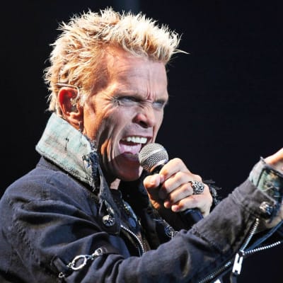 Billy Idol sjunger i en mikrofon och pekar med andra handen.