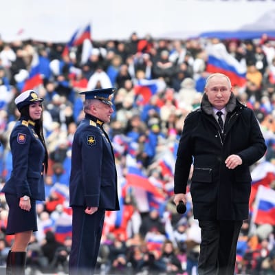 Vladimir Putin kävelee tummassa päällystakissa esiintymislavalla Lužnikin stadionilla.  Hänellä on oikeassa kädessään mikrofoni. Hänestä vasemmalle seisoo kolme univormupukuista ihmistä: kaksi miestä koppalakeissa ja nainen. Taustalla näkyy yleisöä ja Venäjän lippuja katsomossa.