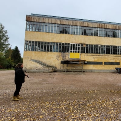 Keltainen, rapistunut suorakaiteen mallinen tehdasrakennus, jota kiertävät kahdessa kerroksessa ikkunarivistöt. Etualalla seisoo nainen selin.