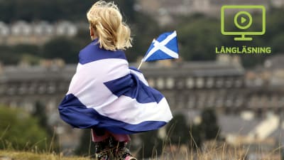 Ett barn tittar över Edinburgh invirat i Skottlands blå-vita flagga och med Skottlands flagga i handen.