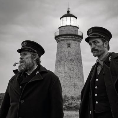 En svartvit bild på Thomas Wake (Willem Dafoe) och Ephraim Winslow (Robert Pattison) står nedanför en fyr och tittar ut mot havet.
