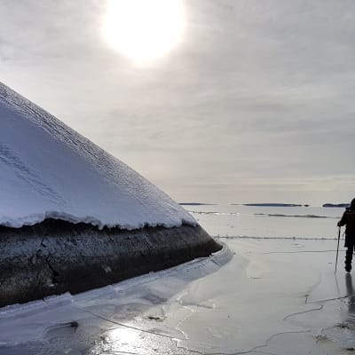 En skridskoåkare står på isen i skärgården, silhuett i motljus.