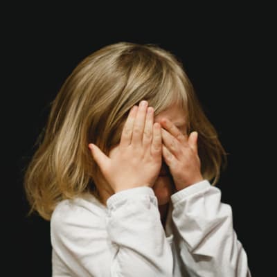Ett barn håller händerna för ögonen och står framör en helsvart bakgrund.