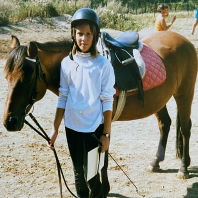 En sadlad brun häst och en ung flicka som står bredvid och håller i tyglarna samtidigt som hon tittar rakt in i kameran. 