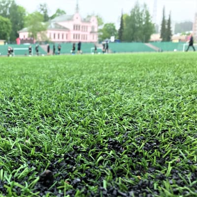 Små svarta kulor ligger bland grässtråna i en konstgräsplan. I bakgrunden syns människor som spelar fotboll i svarta speluniformer.