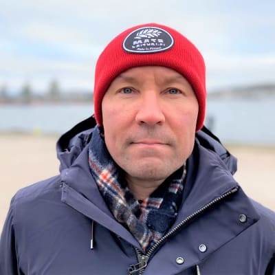 Yle urheilun asiantuntija ja entinen jalkapalloilija Antti Pohja katsoo kameraan. Lähikuvassa on punapipoinen mies järven rannassa.