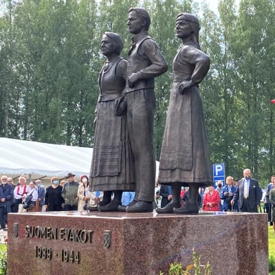 Veistos jossa kolme ihmistä seisoo. Veistos puistossa, ihmisiä ympärillä.