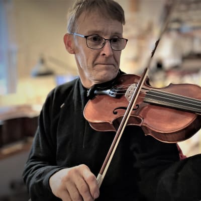 En man med glasögon spelar fiol i en snickarbod. 