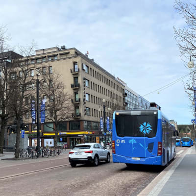 Sininen Lahden paikallisliikenteen bussi parkissa bussipysäkillä, katoksessa odottaa ihmisiä bussia, henkilöauto ohittaa pysähtynyttä bussia. 