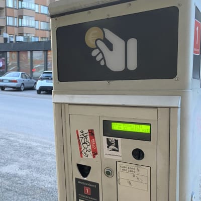 Pysäköintimittari on lähikuvassa lahtelaisen kadun varressa. Kadun toisella puolella on autoja parkissa.
