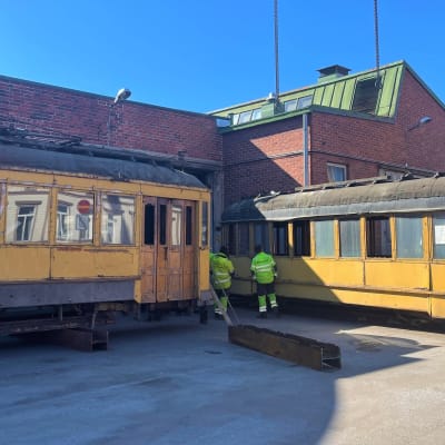 Två gamla, gula spårvagnar står på en gårdsplan framför en röd tegelbyggnad.