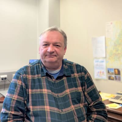 Kärkölän pormestari Markku Koskinen istuu työhuoneessaan, ruutupaita päällään ja katsoo kameraan. Vieressään työpöytä ja seinällä kunnan kartta. 
