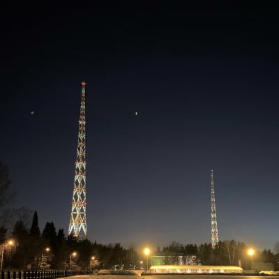 Lahden kaksi radiomastoa iltavalaistuna keskellä talvista maisemaa. Ympärillä radiomäen urheilukentän rakennus ja metsää.