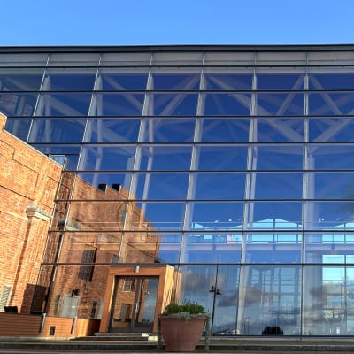 Lähikuvassa on ison rakennuksen lasiseinä ja sen vieressä punatiilinen julkisivu.