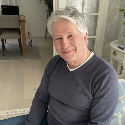 Harmaapaitainen mies istuu sohvalla ja katsoo hymyillen kohti.