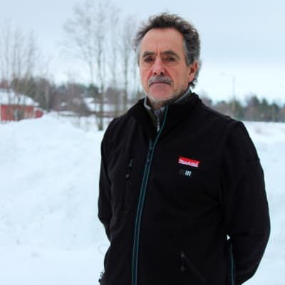 En man i svart jacka och orange byxor står ute i vintermiljö och ser in i kameran med en allvarlig min.