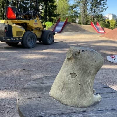 Leikkipuisto. Edustalla näkyy puuveistos, taustalla työmies, työkone ja liukumäkiä.