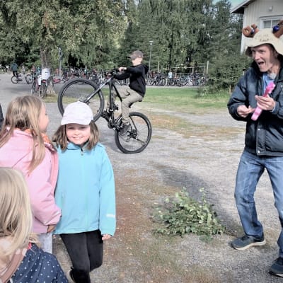 En man med bastuhatt spelar gitarr på en skolgård med ettt gäng barn. 