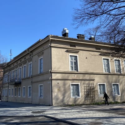 Etualalla on vaalea kivitalo. Taustalla näkyy punainen tiilitalo ja tehtaan piippu. Kadulla rakennuksen edellä kävelee ihminen.