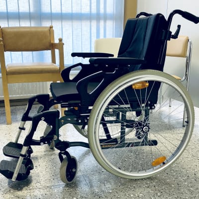 Heinolan terveyskeskuksen aulassa yksinäinen pyörätuoli odottaa käyttäjäänsä. Takana näkyy tuoleja, ikkuna verhon peittämänä.