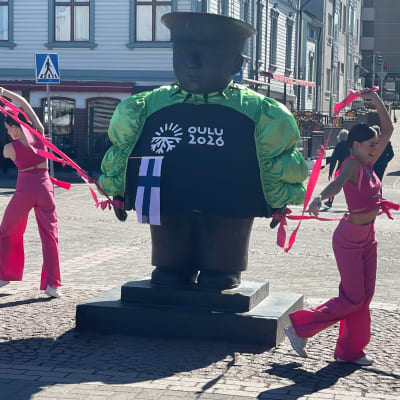 Pinkkiasuiset tanssijat tanssivat Toripolliisi-patsaan ympärillä, patsas on puettu neonvihreään Käärijä-boleroon ja sillä on Suomen lippu kainalossa.